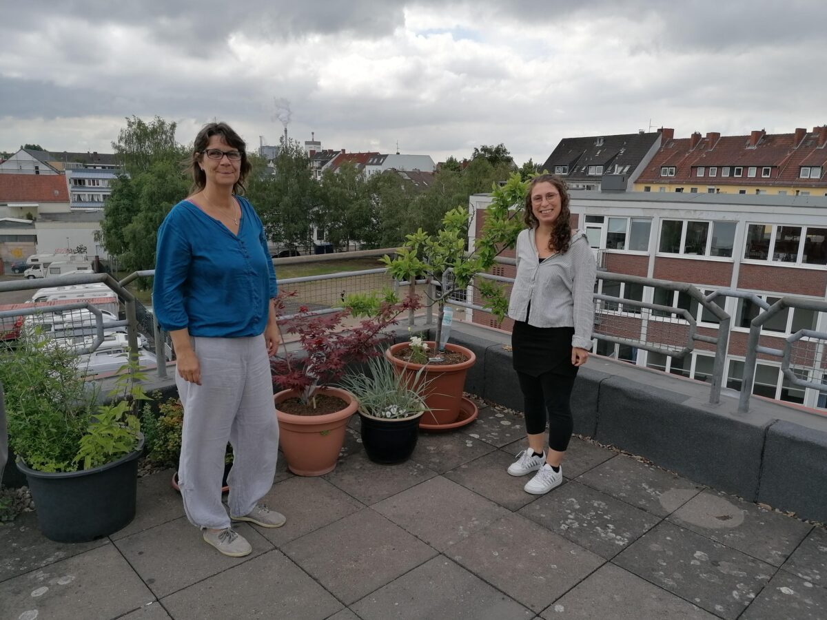 Zwei Frauen stehen auf einer Dachterasse. Im Hintergrund ein Häuserpanorama.